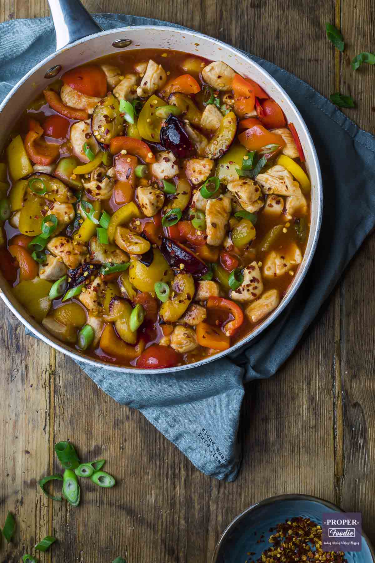 Sweet and sour chicken in a large pan topped with spring onions and chilli flakes.