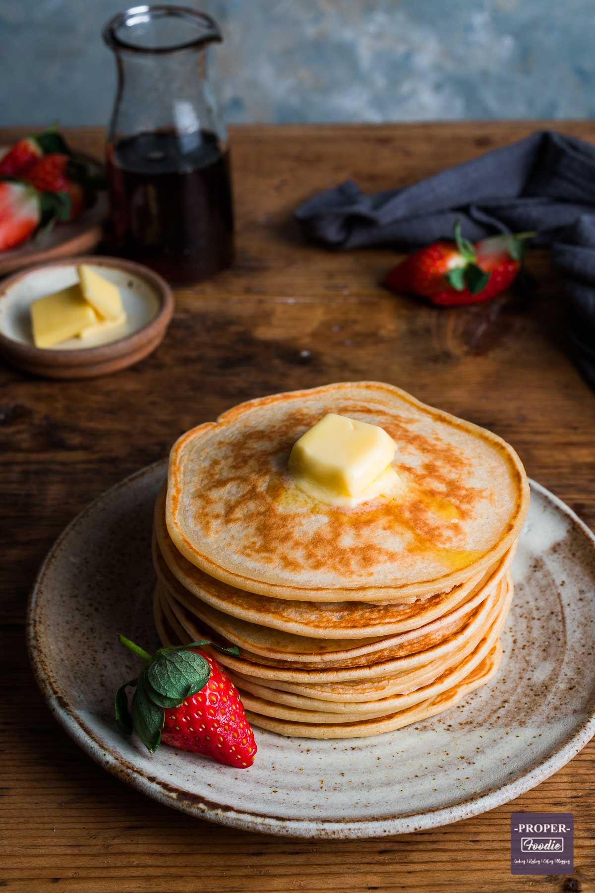 A stack of pancakes on a small plate with butter melting on the top and a small glass jug of syrup in the background.