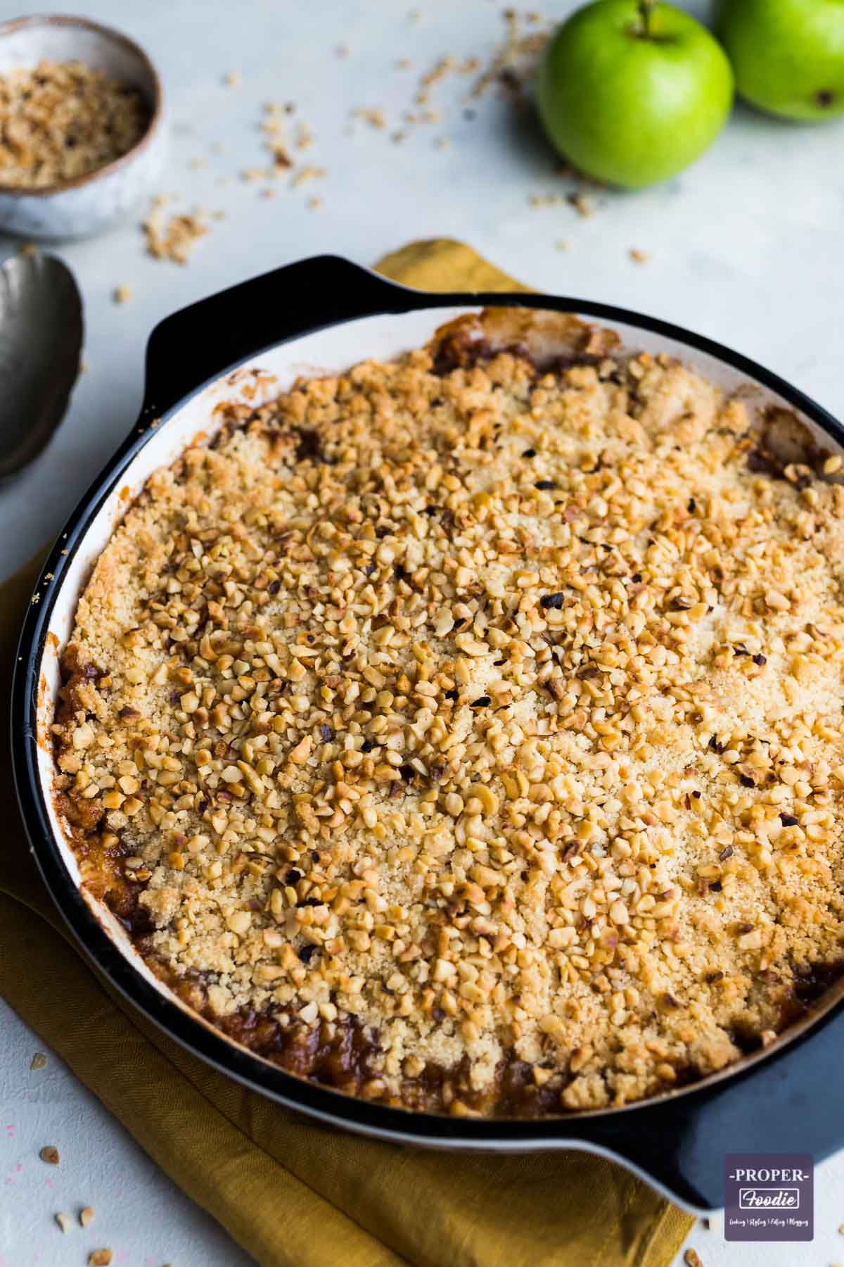 Round baking dish filled with apple crumble and topped with chopped hazelnuts, and baking apples the background.