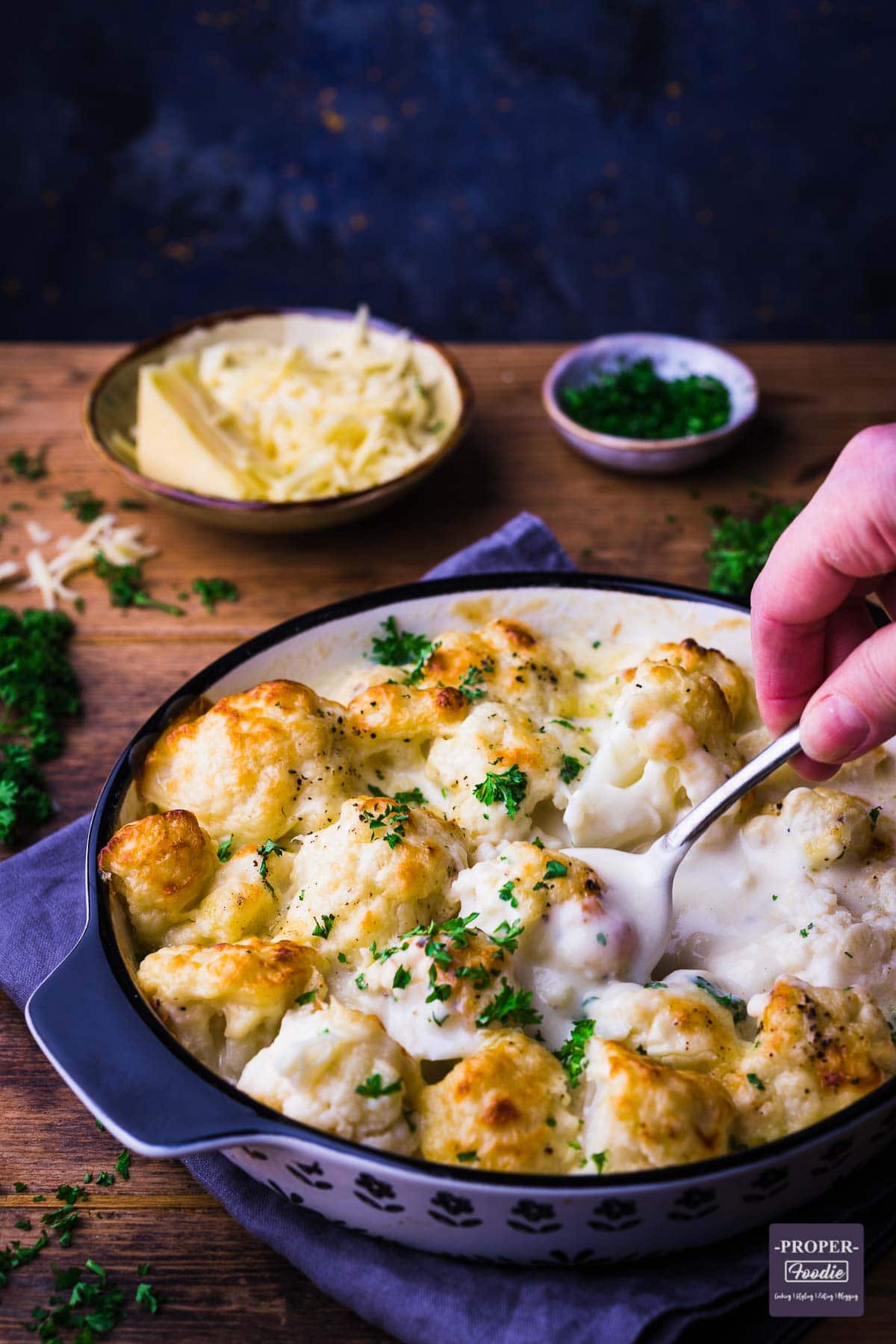 Cauliflower presented in a shallow dish with a creamy sauce and topped with baked on cheese and freshly chopped parsley.