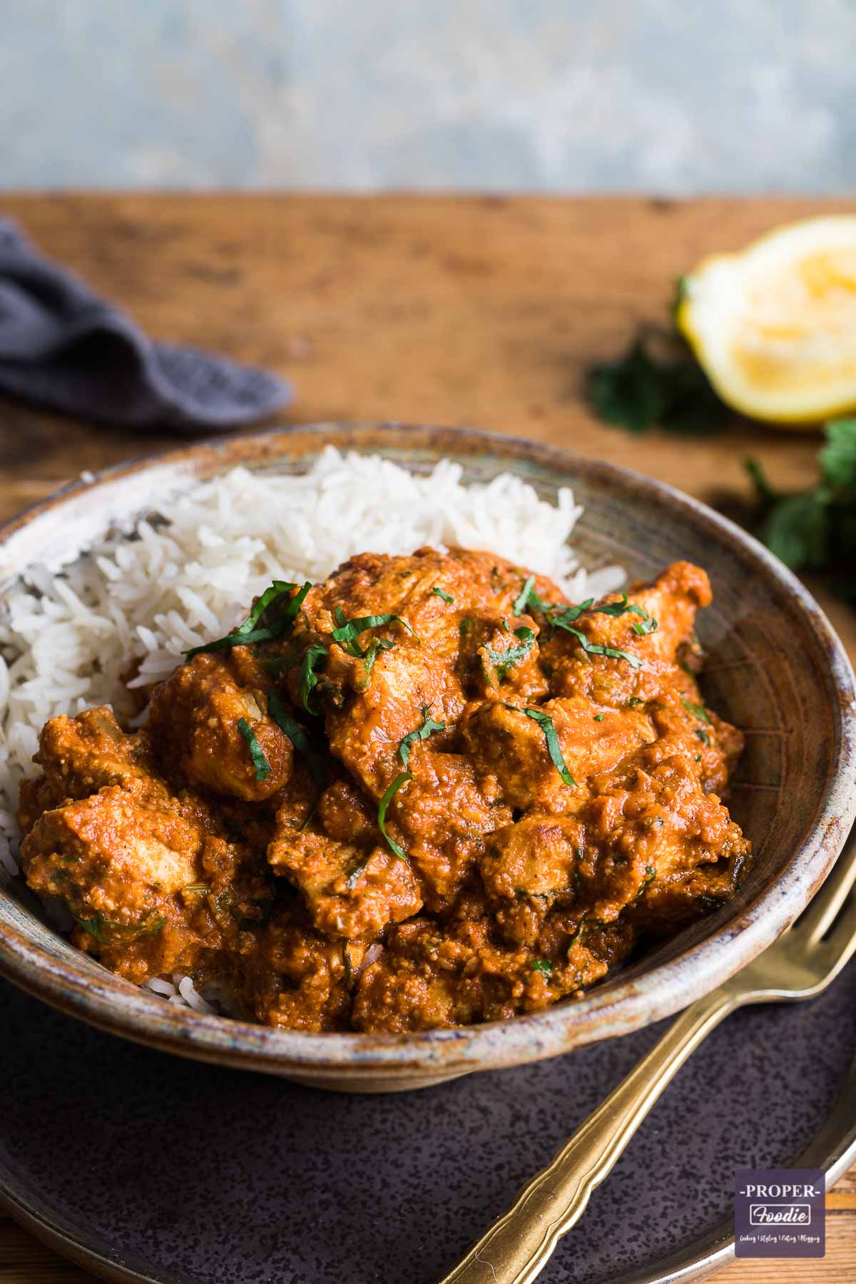 Chicken tikka masala curry and rice in a bowl sat on a plate with a gold fork at the side and half a squeezed lemon in the background.