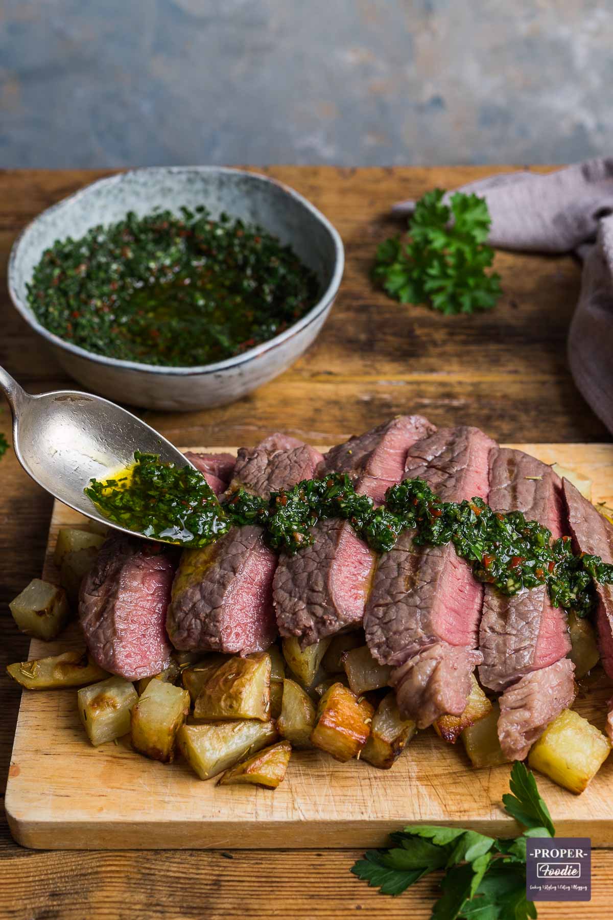 Slices of medium-rare steak positioned over a pile of cubed rosemary potatoes with a herb and chilli dressing being spooned over the top.