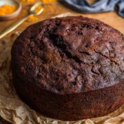 Fruit Christmas cake without icing sitting on brown paper, on a bronze cooling rack, with orange zest and a gold spoon in background.