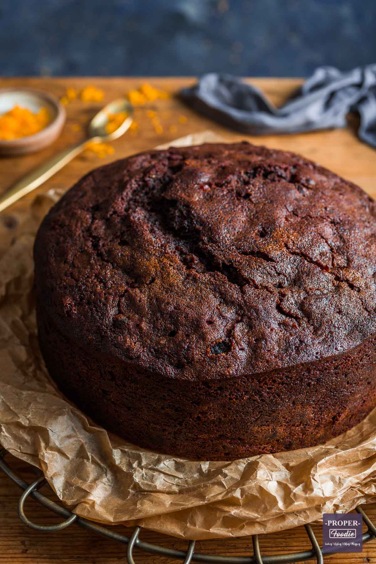 Fruit Christmas cake without icing sitting on brown paper, on a bronze cooling rack, with orange zest and a gold spoon in background.