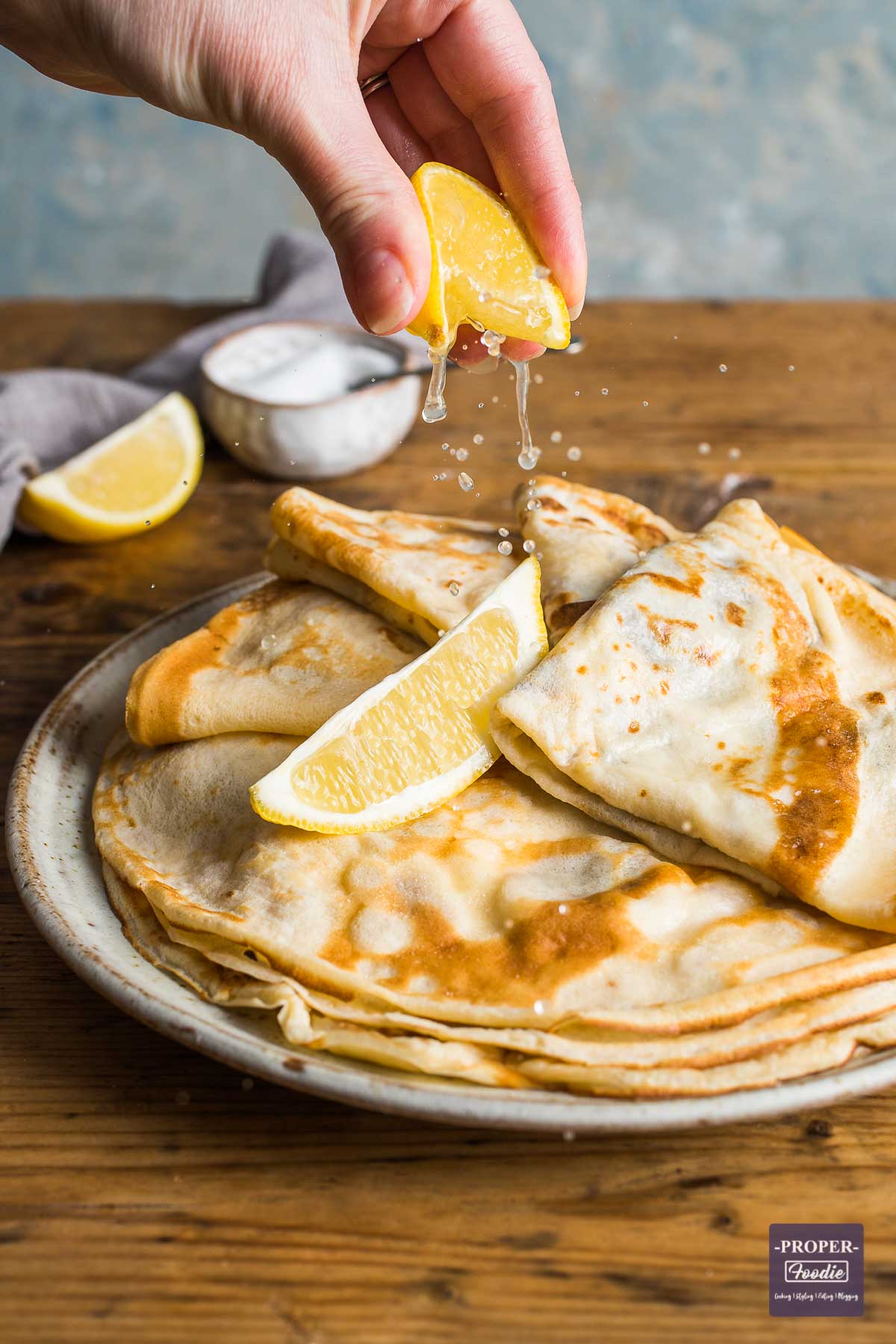 A plate of this flat pancakes stacked with the top four folded twice topped with a lemon wedge and a second wedge being squeezed over the top.