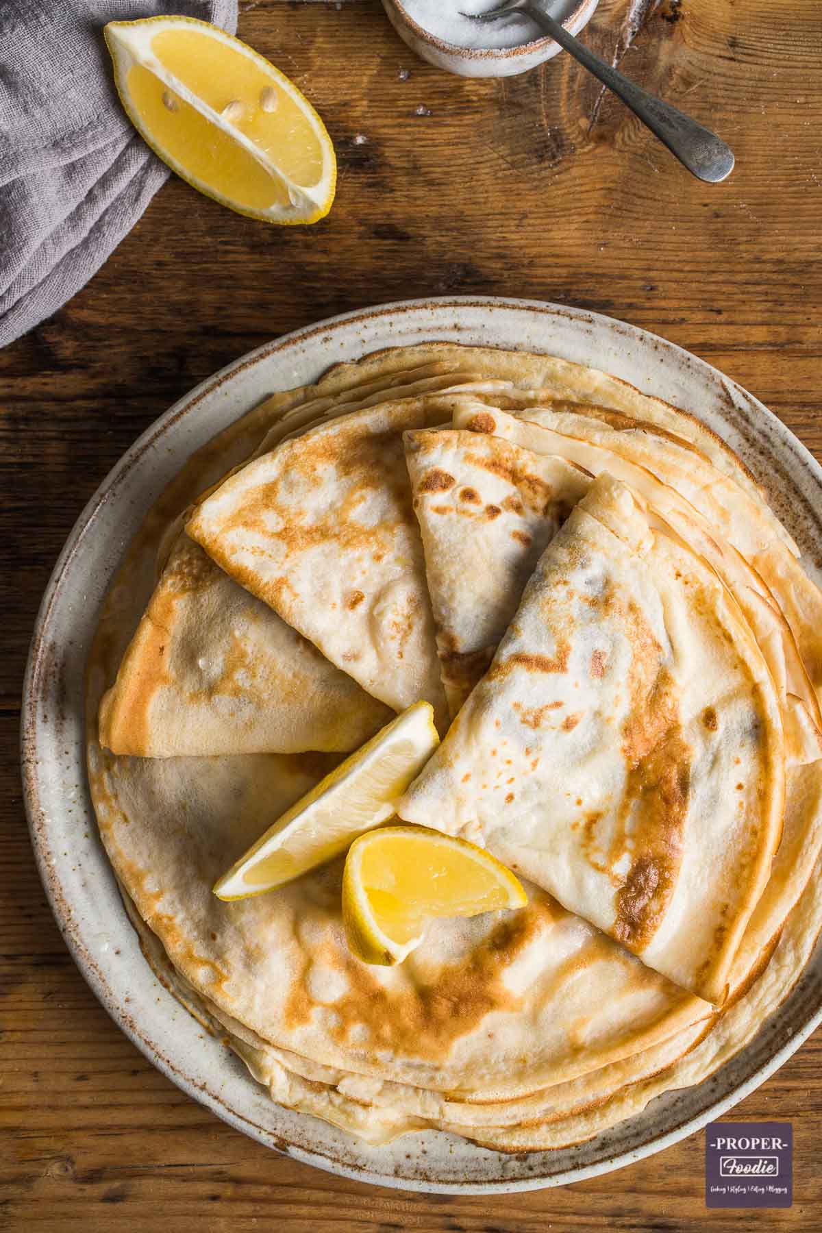 Plate of thin, flat English pancakes viewed from above with the top 4 folded into quarters ad 2 lemon wedges placed on top.