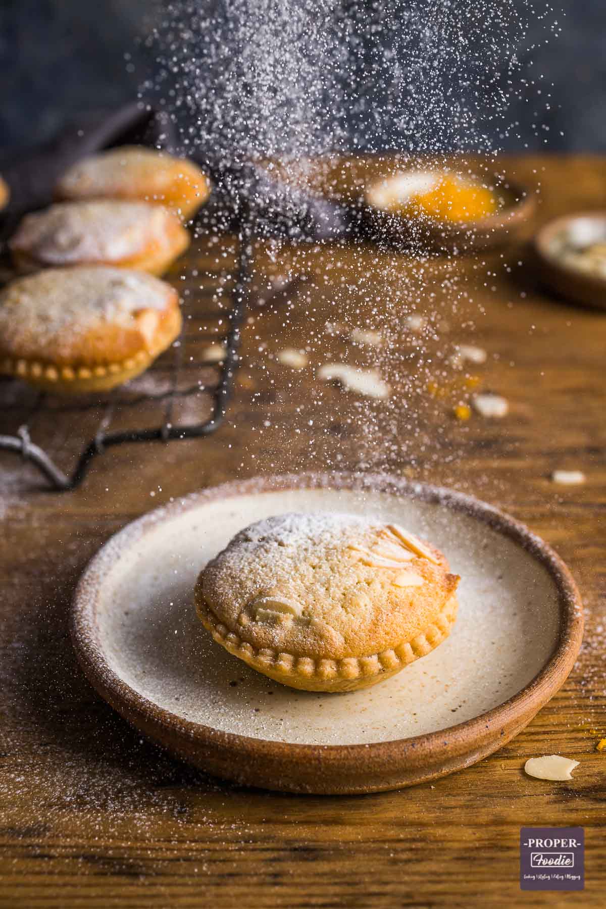 Single mince pie on a plate with a frangipane topping and sprinkles of icing sugar falling down on top.