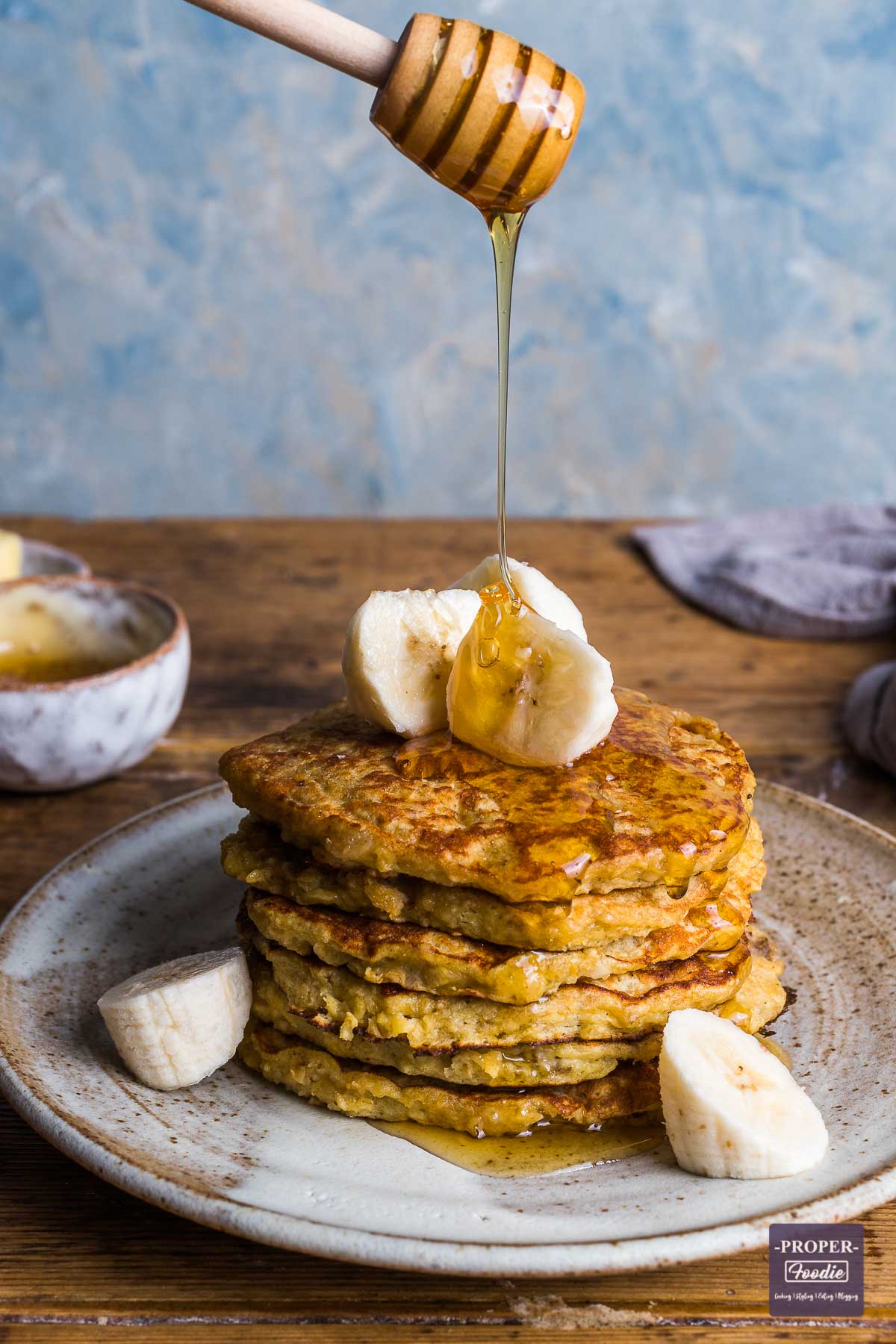 A stack of banana pancakes on a plate topped with slices of banana and a line of honey being poured over from honey dipper.