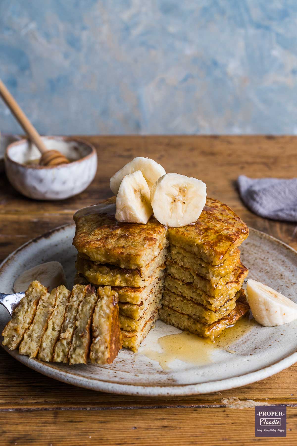 Stack of banana pancakes topped with sliced bananas, and with a wedge skewered and cut out from one side of the pancake stack and put to the side.