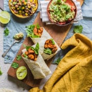 three chicken wraps on a board viewed from above with half a lime squeezed at the side and small bowls of bean salad and guacamole above