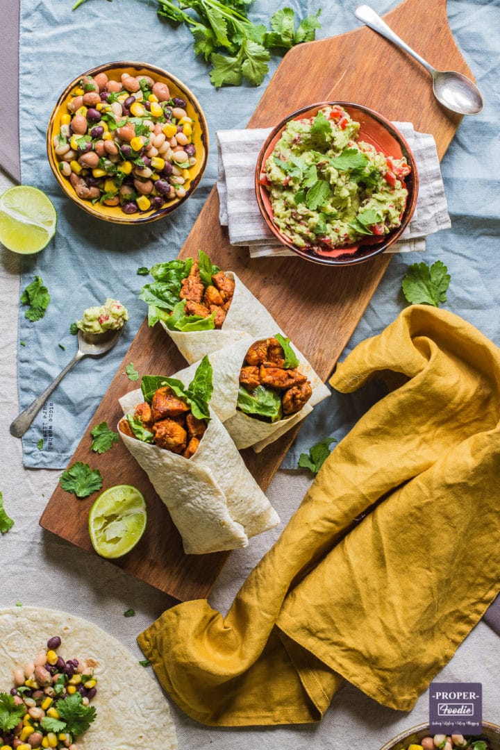 three wraps with chicken and lettuce viewed from above with half a lime squeezed at the side and small bowls of bean salad and guacamole above