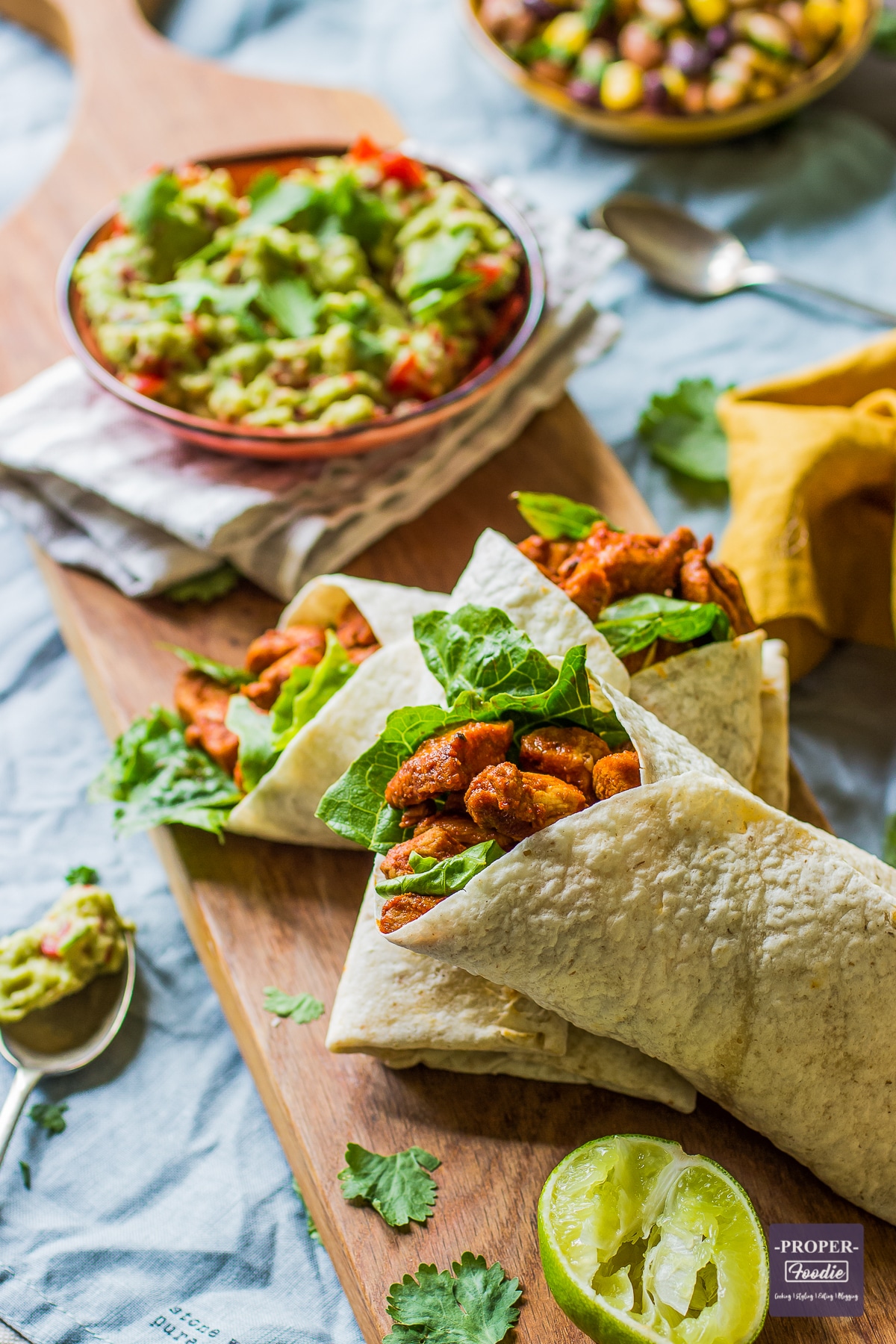 three chicken wraps presented on a long slim board with a bowl of guacamole and a squeezed lime