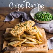 Homemade fish and chips on a wooden board with a bowl of garden peas in the background