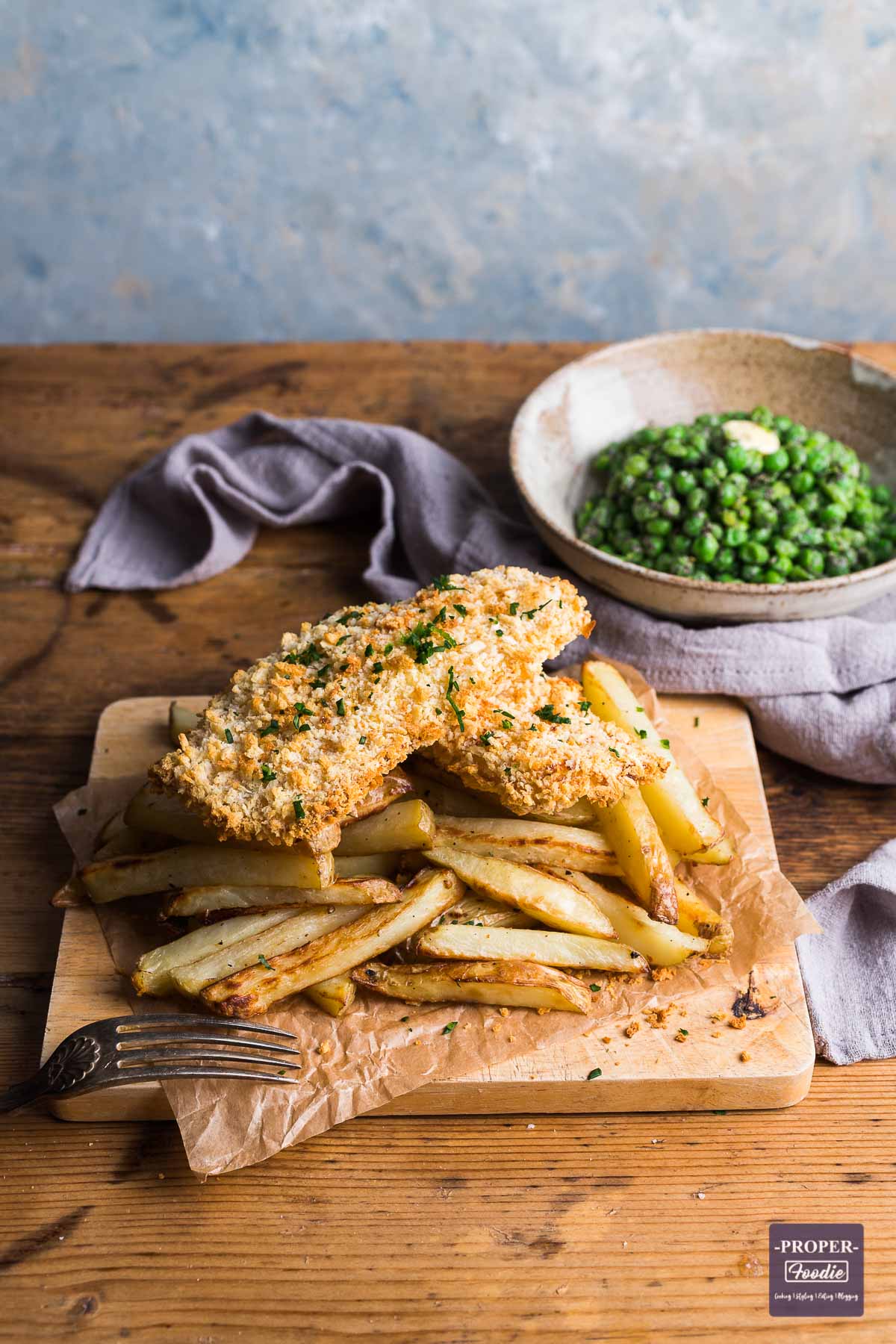 Bread crumbed fish fillets sat on a pile of homemade chips with a bowl of peas in the background topped with melting butter