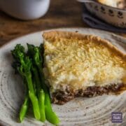 A slice of lamb and mint pie with a pastry base and mash potato topping, served on a plate with tender stem broccoli.