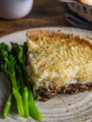 A slice of lamb and mint pie with a pastry base and mash potato topping, served on a plate with tender stem broccoli.