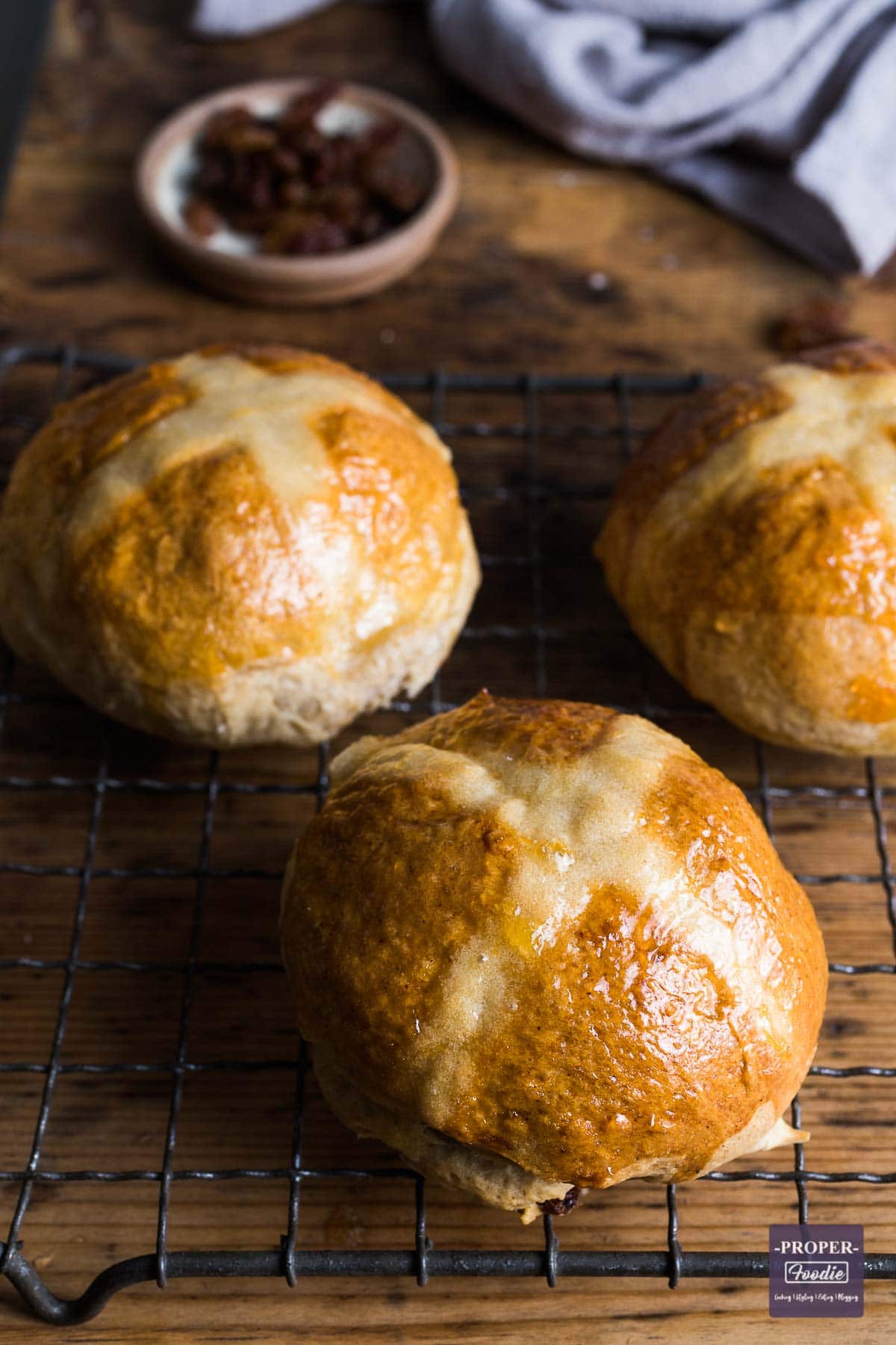 three homemade hot cross buns on a cooli rack with a small dish of sultanas in the background