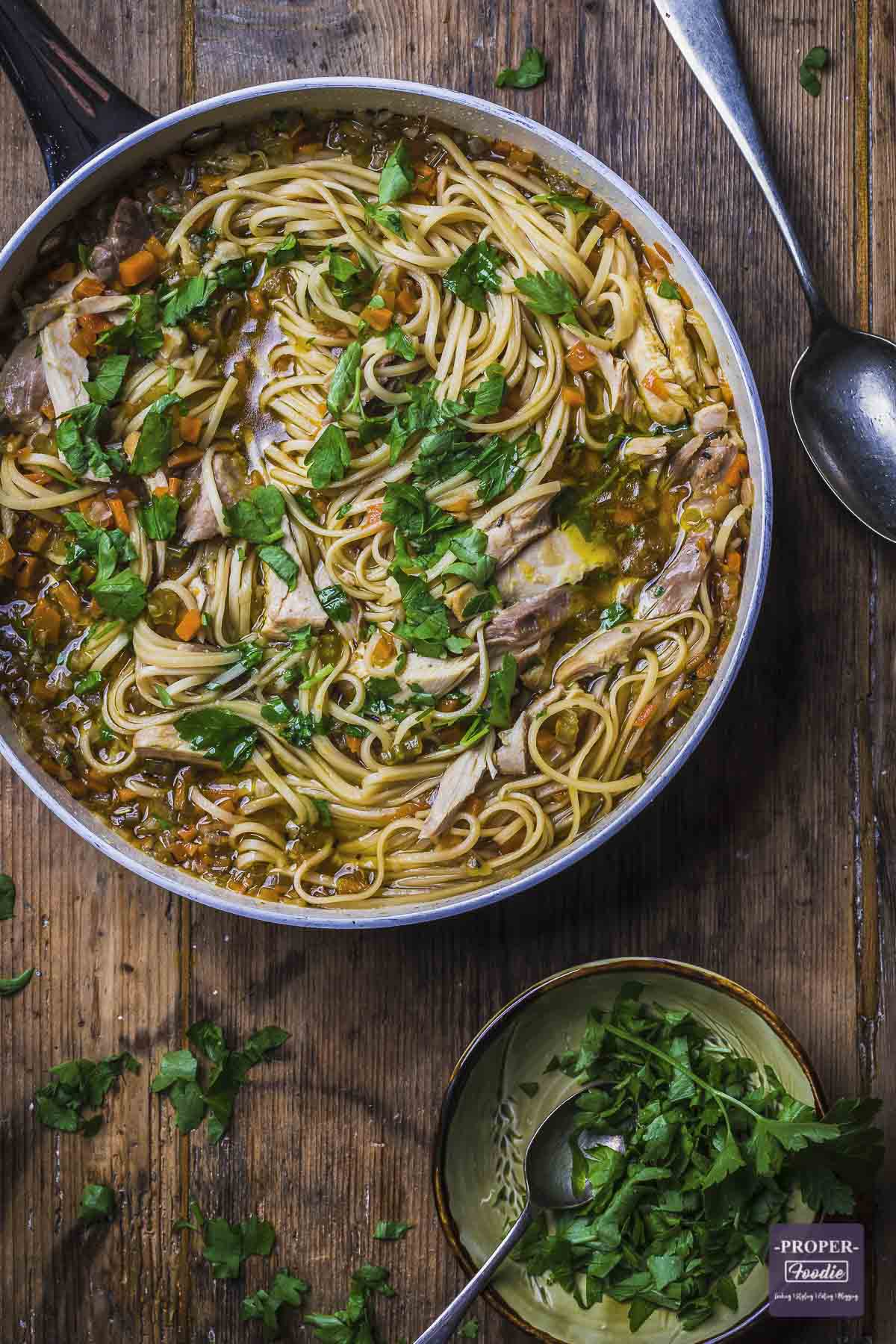 A large pan of chicken noodle soup with chopped vegetables and fresh parsley.