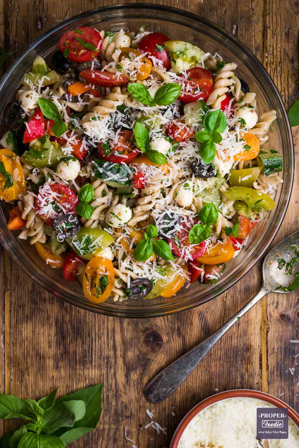 Large bowl filled with cooked fusilli pasta, colourful tomatoes, cucumber, mozzarella pearls and topped with basil leaves and grated pecorino.