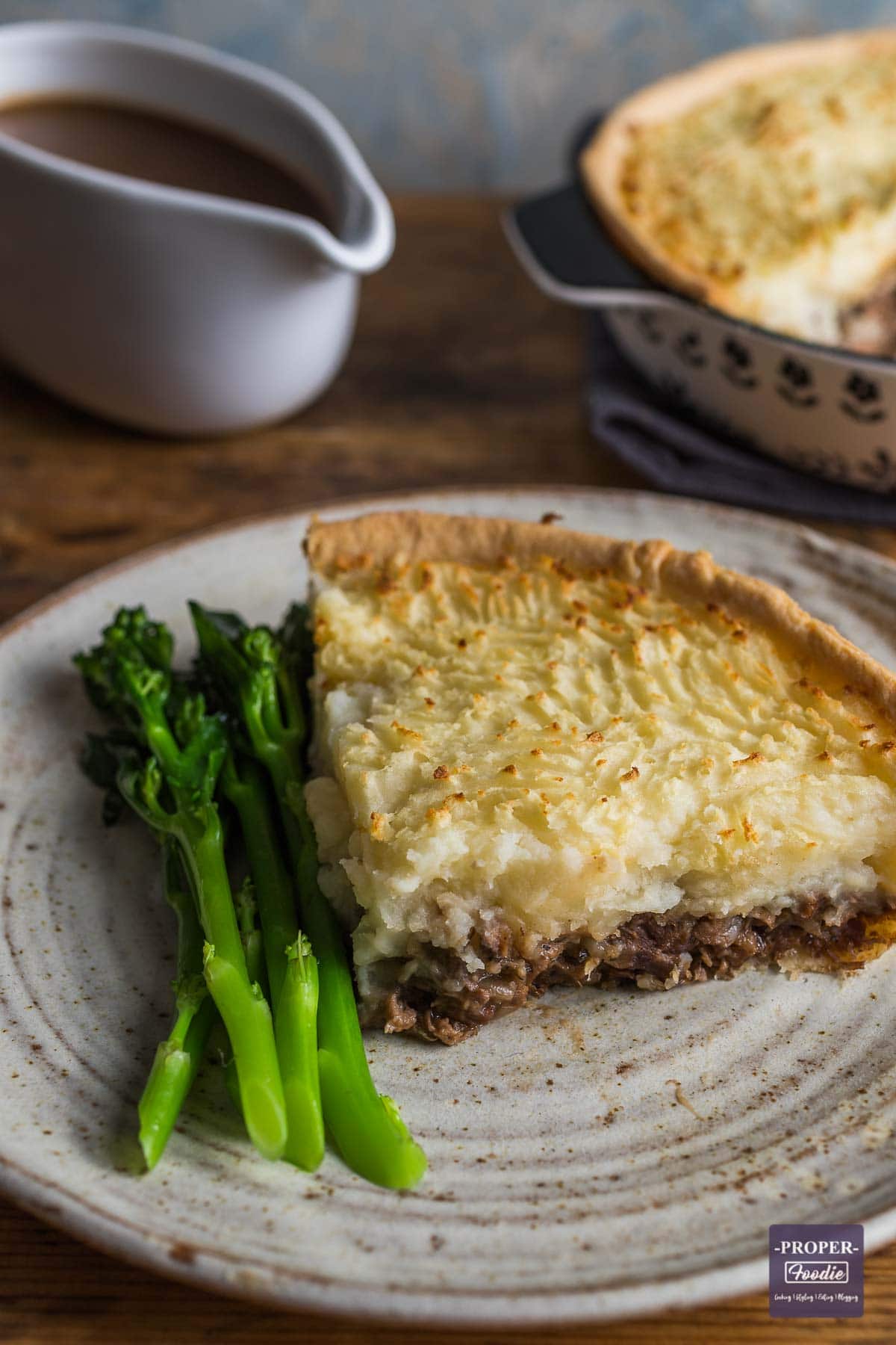 A slice of lamb and mint pie with a pastry base and mash potato topping, served on a plate with tender stem broccoli.