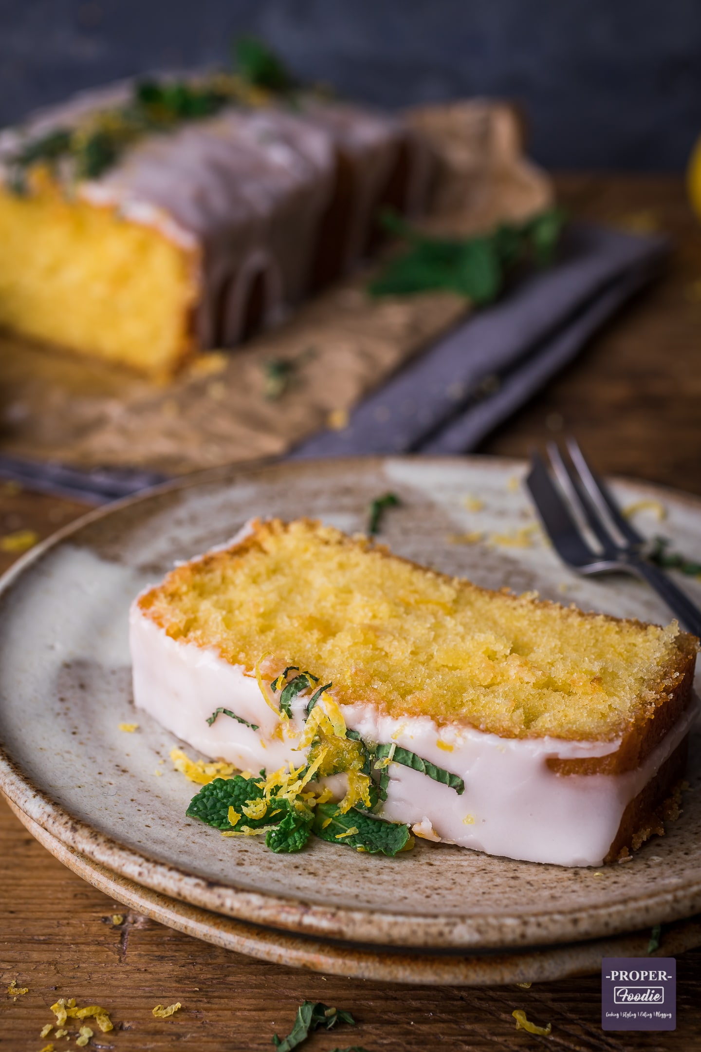 Slice of lemon cake on a plate with the rest of the cake in the background