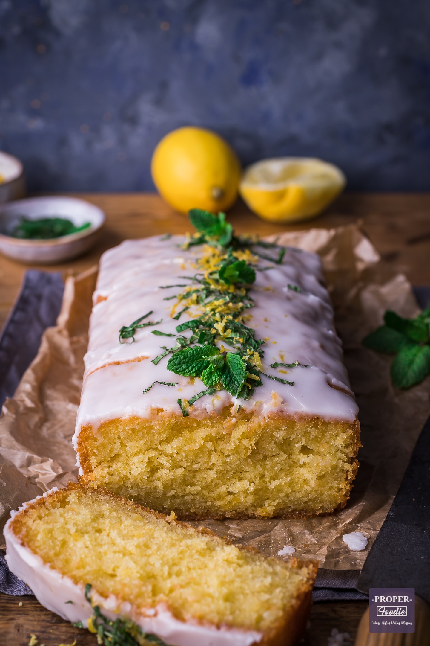 lemon drizzle cake with one piece sliced off at the front and lemons and fresh mint in the background