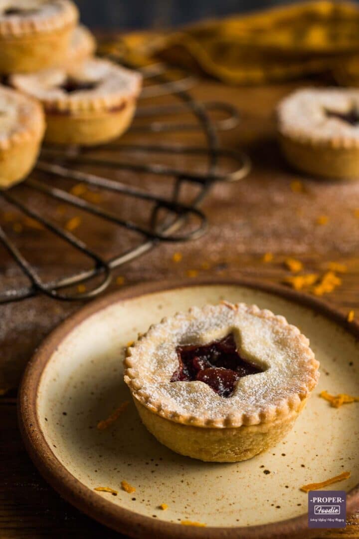 A single mince pie on a small plate with a stack of mince pies on a cooling rack in the background.