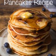 Fluffy blueberry pancakes stacked on a small plate with golden syrup being poured over the top.