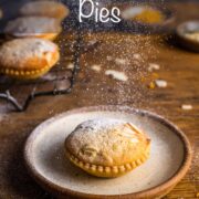 Single mince pie on a plate with a frangipane topping and sprinkles of icing sugar falling down on top. Overlay text: Frangipane Mince Pies.