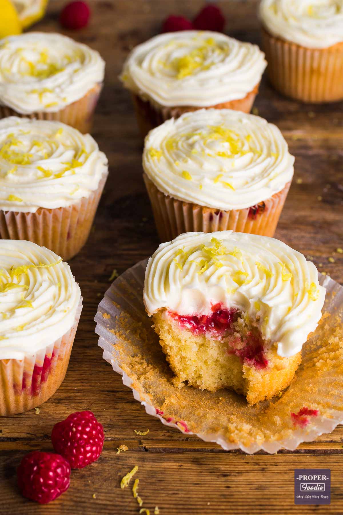 Raspberry muffin topped with lemon butter cream sitting on a muffin case with the sides pulled away and surrounded a few other muffins.