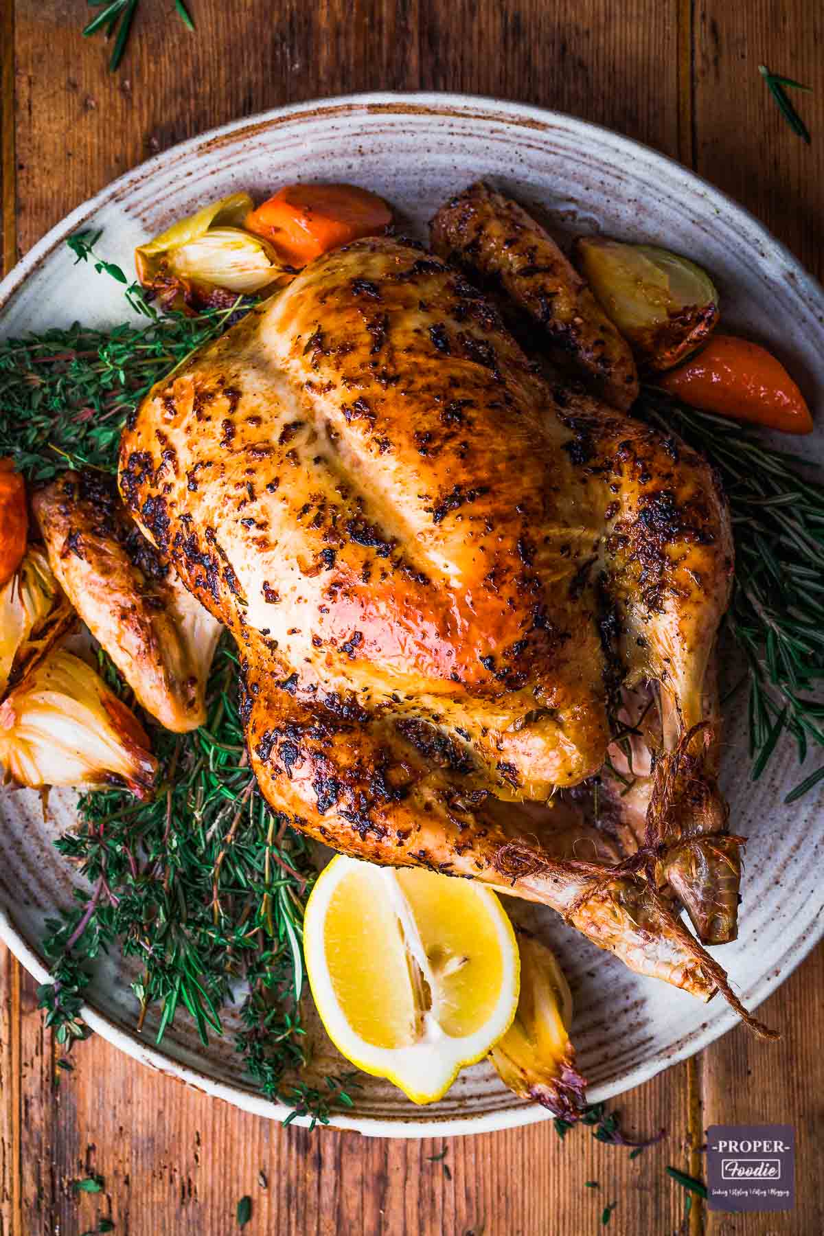 whole chicken roasted and presented on a large plate with cooked carrots and onions and fresh herbs.