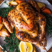 whole chicken roasted and presented on a large plate with cooked carrots and onions and fresh herbs.