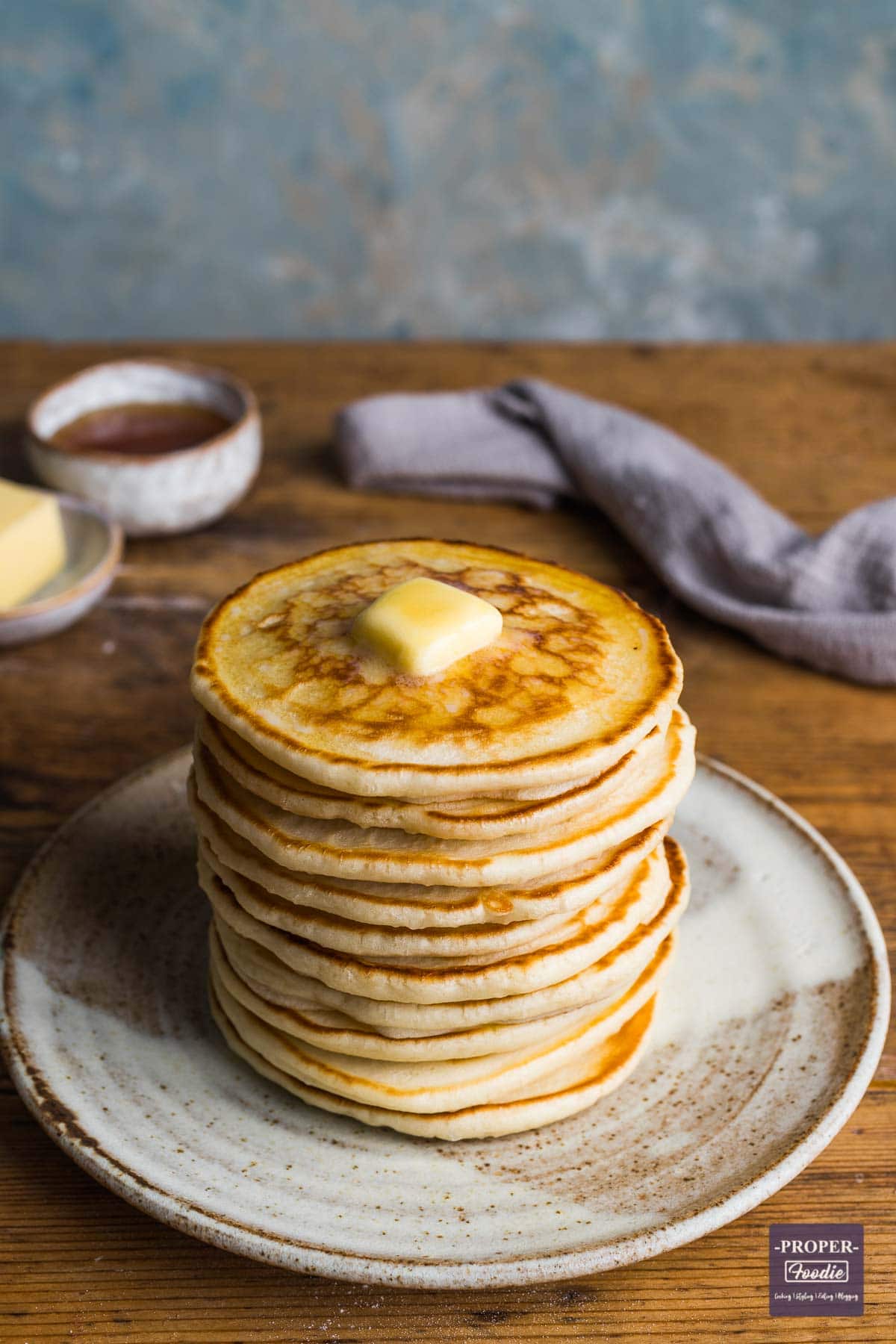 A tall stack of scotch pancakes on a plate with melted butter on top ad a small dish on honey in the background.