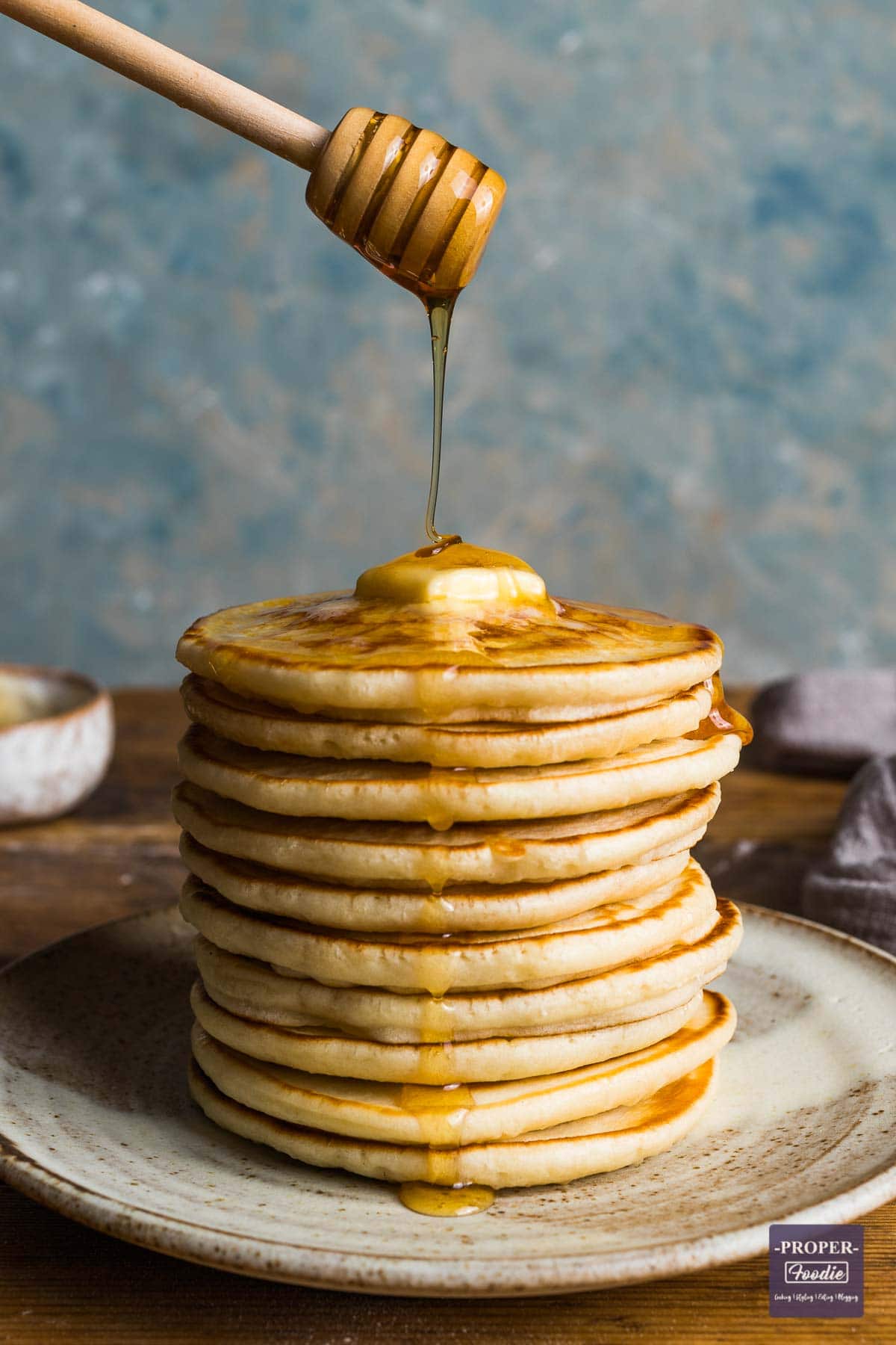 A tall stack of scotch pancakes on a plate with melted butter on top and honey being drizzled over with a wooden honey dipper.