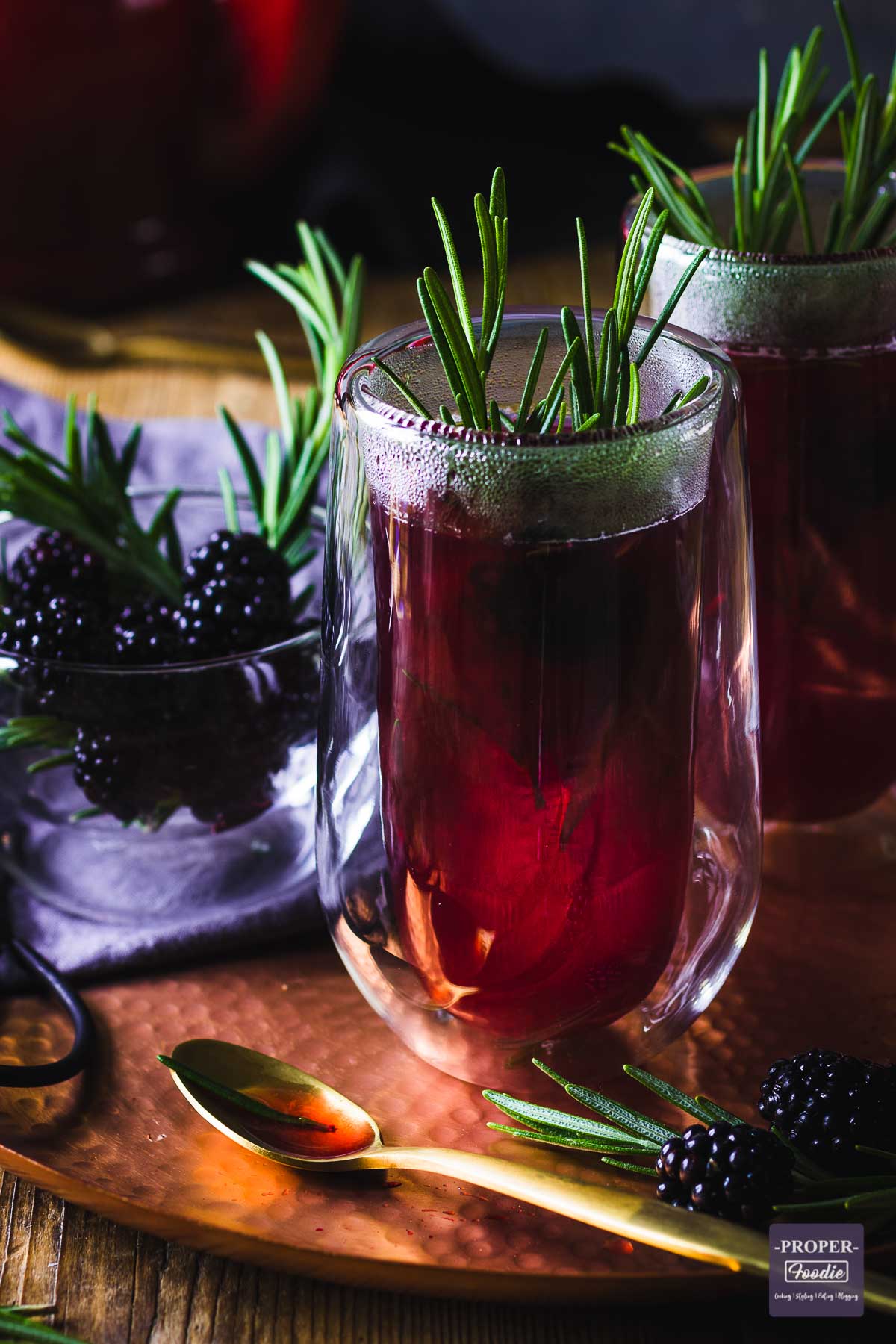 a purple and steaming, double walled glass of sloe gin hot toddy decorated with a sprig of rosemary and a second glass in the background.