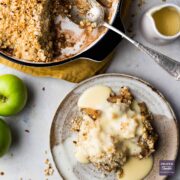 A portion of apply crumble on a small plate with custard drizzled over and the rest of the crumble in a larger dish with a spoon.