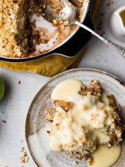 A portion of apply crumble on a small plate with custard drizzled over and the rest of the crumble in a larger dish with a spoon.