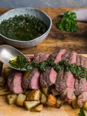 Slices of medium-rare steak positioned over a pile of cubed rosemary potatoes with a herb and chilli dressing being spooned over the top.