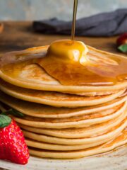 A stack of American pancakes with a piece of butter melting on the top and syrup poured over and drizzling down one side.