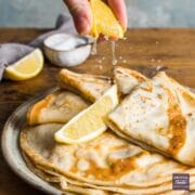 A plate of this flat pancakes stacked with the top four folded twice topped with a lemon wedge and a second wedge being squeezed over the top.