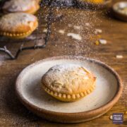 Single mince pie on a plate with a frangipane topping and sprinkles of icing sugar falling down on top.