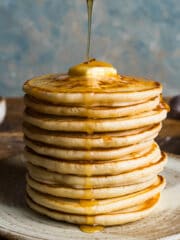 A tall stack of scotch pancakes on a plate with melted butter on top and honey being drizzled over with a wooden honey dipper.