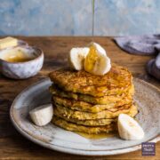 A stack of banana pancakes on a plate topped with slices of banana and a line of honey being poured over from above.