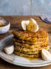 A stack of banana pancakes on a plate topped with slices of banana and a line of honey being poured over from above.