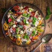 Large bowl filled with cooked fusilli pasta, colourful tomatoes, cucumber, mozzarella pearls and topped with basil leaves and grated pecorino.