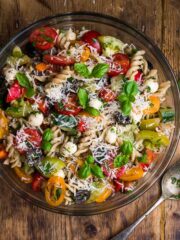 Large bowl filled with cooked fusilli pasta, colourful tomatoes, cucumber, mozzarella pearls and topped with basil leaves and grated pecorino.