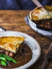 A slice of steak and ale pie on a plate with gravy and tender-stem broccoli with pie dish in the background.