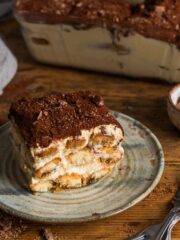 A small, square piece of tiramisu on a plate with the side view showing layers of sponge fingers and creamy mascarpone. The top is dusted with cocoa powder and a large dish of tiramisu is in the background.