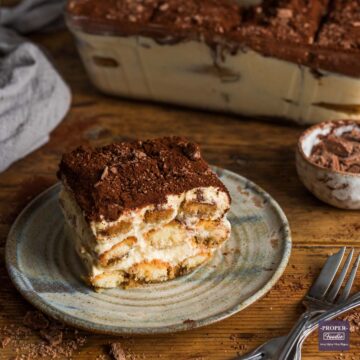 A small, square piece of tiramisu on a plate with the side view showing layers of sponge fingers and creamy mascarpone. The top is dusted with cocoa powder and a large dish of tiramisu is in the background.