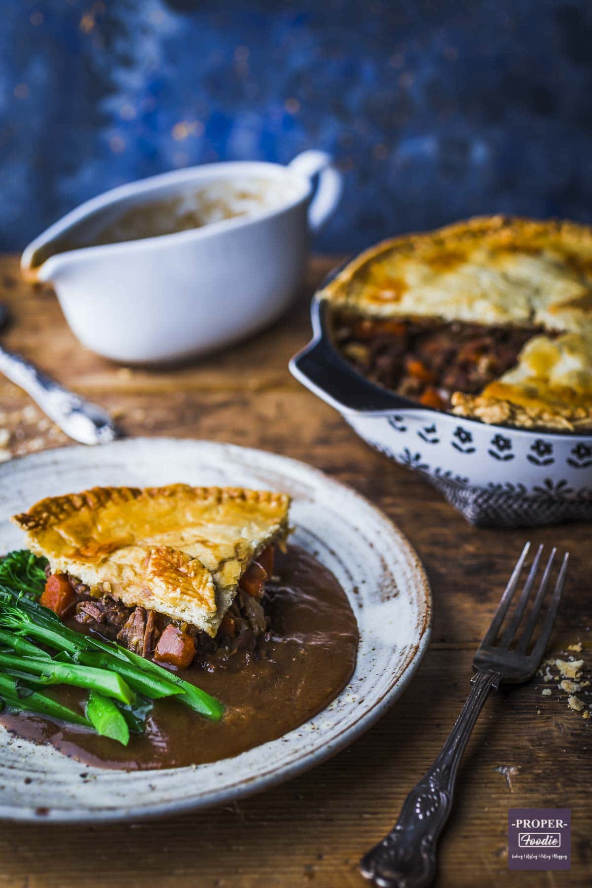 Homemade steak and ale pie served on a plate with pie dish and gravy boat in the background.